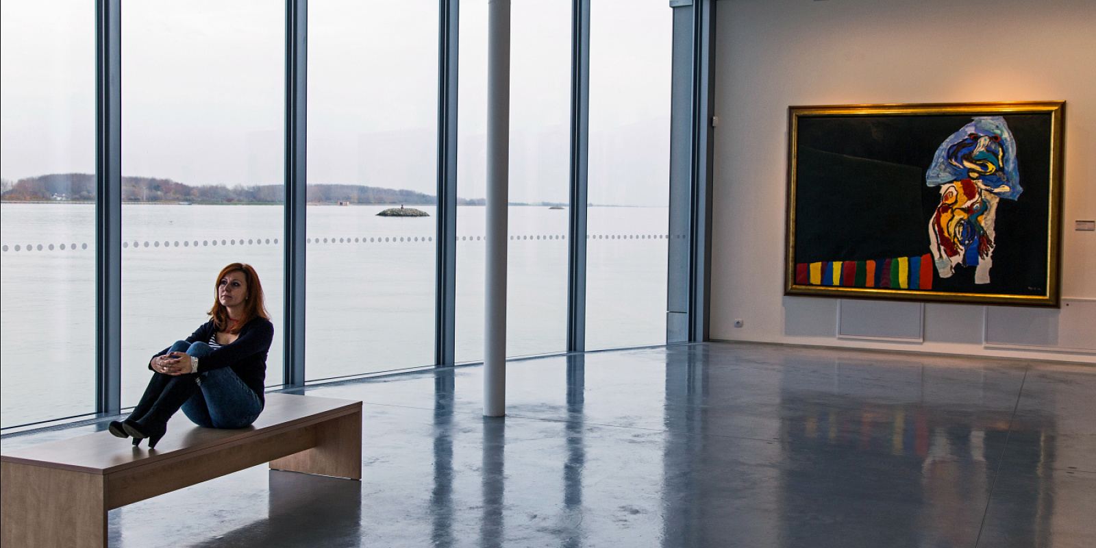 Woman sitting at Danubiana Museum, viewing the Danube through large windows.