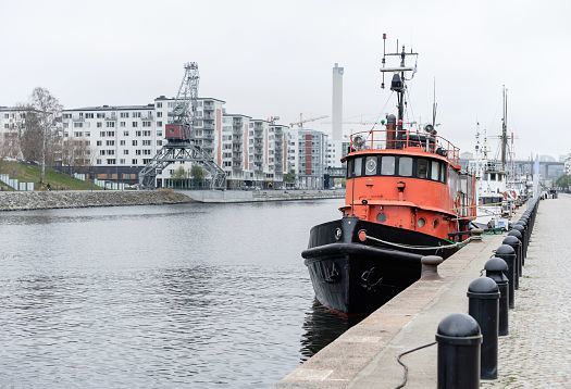 A red pilot boat is docked in the harbor of Stockholm, Sweden, with residential buildings and a crane in the background.
