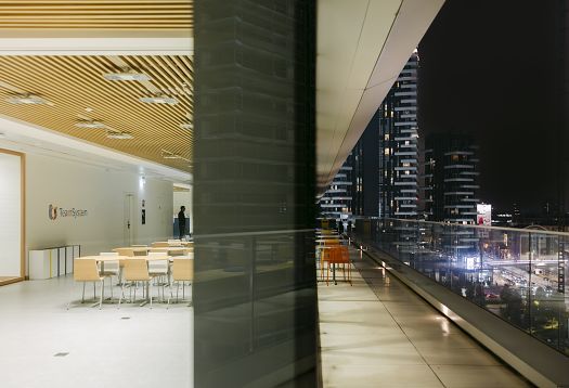 Interior of a modern office building with a slatted timber ceiling and bright seating areas on the left; on the right, a wide covered terrace with a glass balustrade and views of illuminated skyscrapers at night.