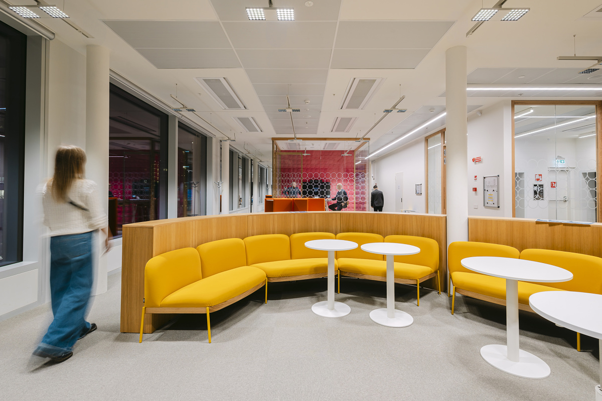 Modern office area with a curved yellow bench and white round tables in the foreground. In the background, a glass-walled meeting room with red wall paneling can be seen, with people moving through the room.
