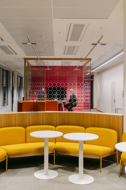 Modern office area with yellow benches and white round tables in the foreground. Behind them is a glass-walled meeting room with red wall paneling and two people seated at a table.