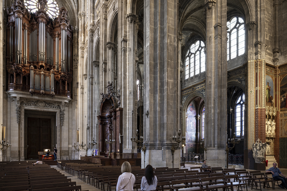 Interior of a large historic church showing tall stone columns, vaulted ceilings, and an ornate wooden pipe organ mounted above a large doorway. Rows of wooden pews fill the foreground, and two visitors stand near the center, looking toward the architectural details and artworks on the right wall.