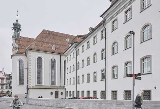 View of a historic building façade with light-colored walls, tall windows, and an adjoining church; people on a cobbled street in the foreground.